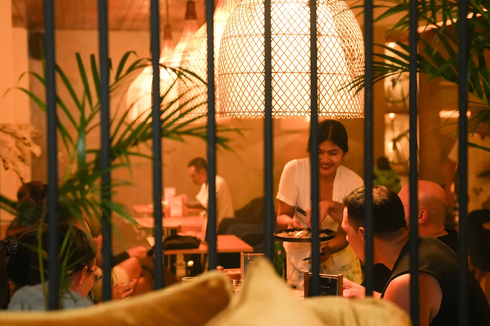 A candid view from a sofa seat through decorative vertical bars into a lively and atmospheric restaurant in Ubud. Diners are enjoying their top dinner in Ubud under warm, large woven pendant lights, with a smiling server tending to a table. The environmen