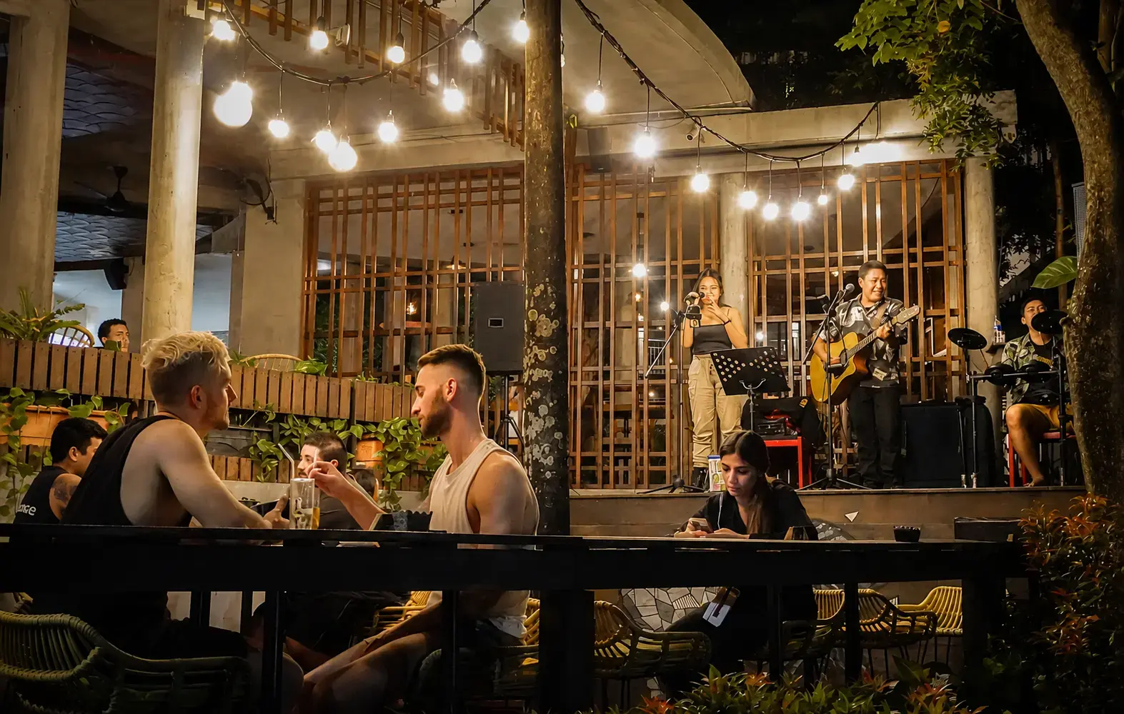 Outdoor seating area and live music stage at Dua Sisi Kitchen & Bar, Grandma's Plus Hotel, Seminyak, with string lights create a vibrant and inviting atmosphere perfect for casual dining.