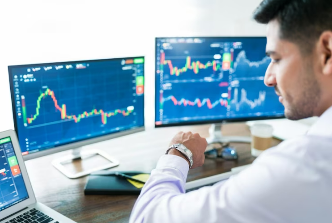 Profiled man wearing a white shirt and looking at his wristwatch, in front of three monitors displaying price charts and crypto technical analysis