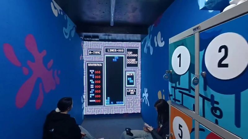 Комната с яркими росписями на стенах: на LED-стене идёт игра «Тетрис», двое посетителей сидят перед ней с геймпадами