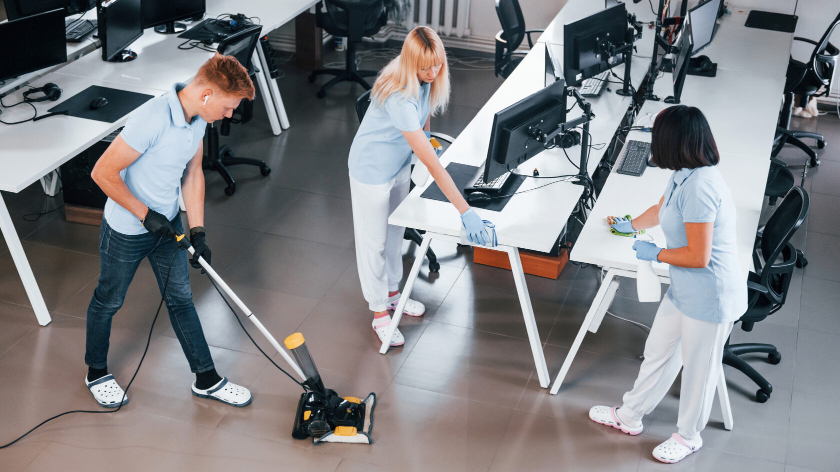 Три клинера в униформе работают в офисе. Один использует пылесос для уборки пола, а два других протирают столы и компьютеры. Все клинеры работают в перчатках и защитной одежде.