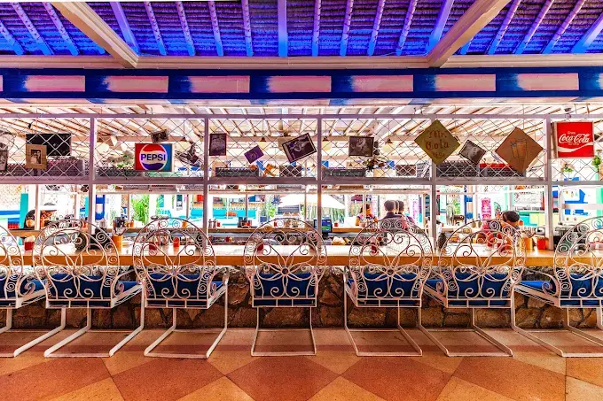 The vibrant, retro-neon bar counter and white wrought-iron seating at Motel Mexicola, known for its high-energy Mexican party atmosphere in Seminyak.