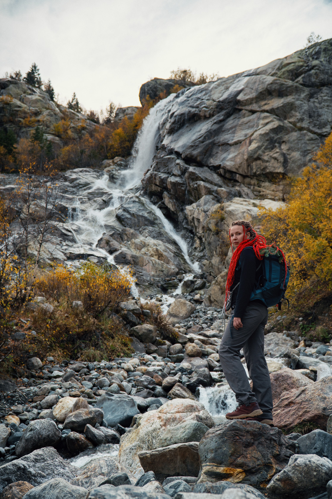 Водопад Алибек. Осень в горном лесу, горный водопад стремится вниз по скалам. Девушка с красными дредами стоит напротив водопада. Золотая осень в горах. Посёлок Домбай.