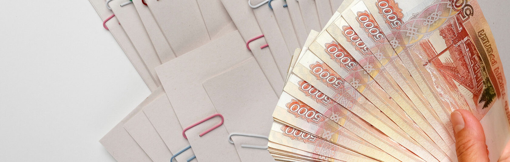 Рука держит веер банкнот рублей рядом с папками документов