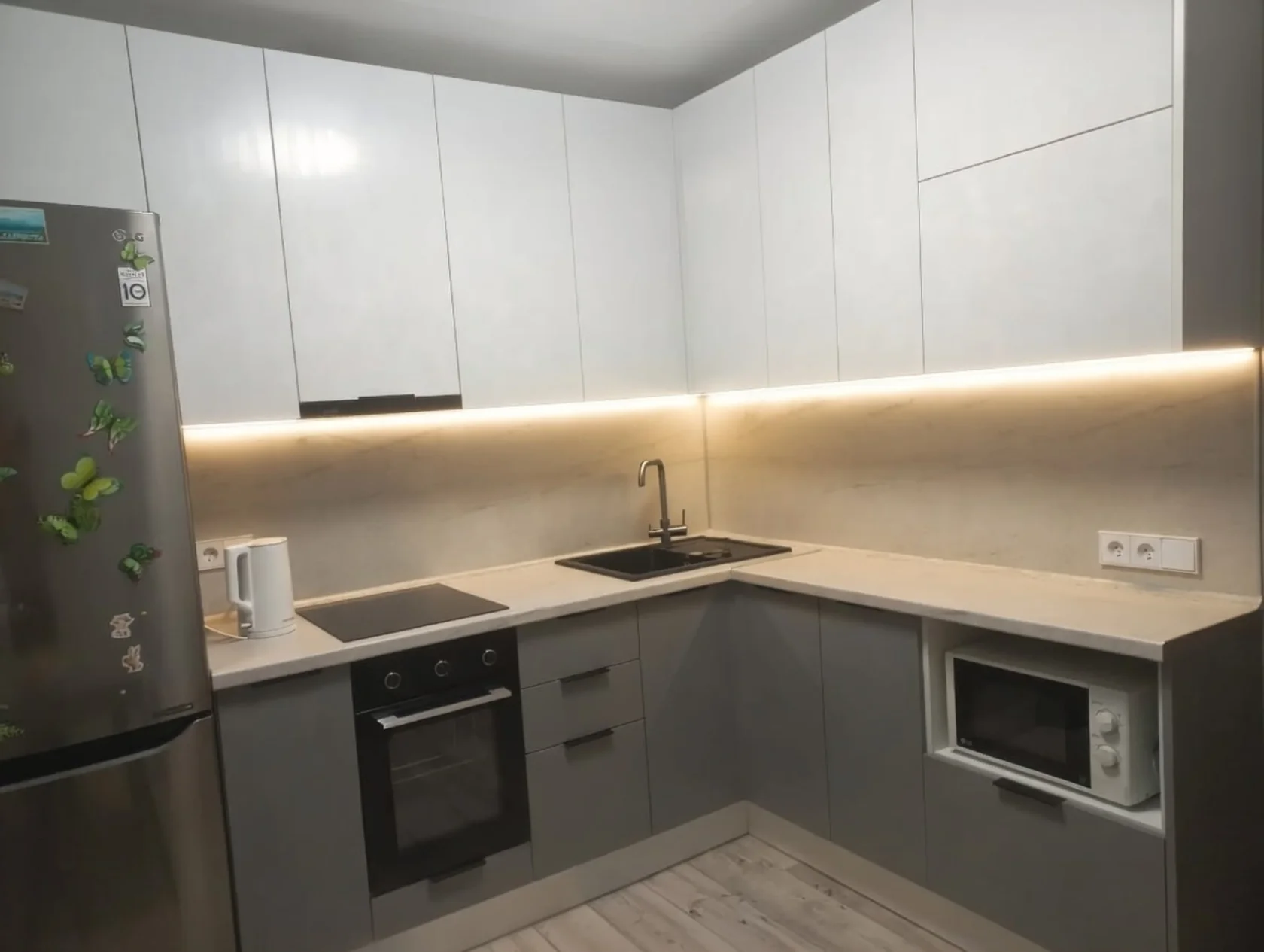 Угловая кухня на заказ в светлых тонах: белые верхние фасады, серые нижние шкафы, мраморная столешница, черная мойка, индукционная варочная панель, встроенный духовой шкаф и микроволновка, LED-подсветка под верхними шкафами Современная маленькая кухня в серо-белой гамме с магнитиками на холодильнике, подсветкой рабочей зоны и техникой Bosch / LG Готовая угловая кухня эконом-класса: белые глянцевые фасады, серая нижняя часть, столешница под светлый мрамор, черная врезная мойка и варочная поверхность