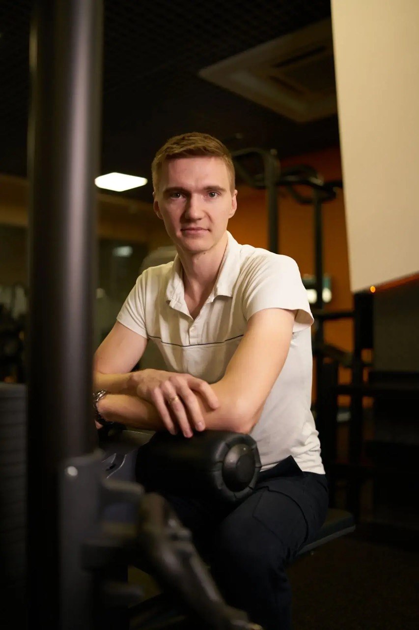 Артём Карпекин, персональный тренер Orange Fitness категории «элит» и фитнес-нутрициолог, в белом поло у тренажёра