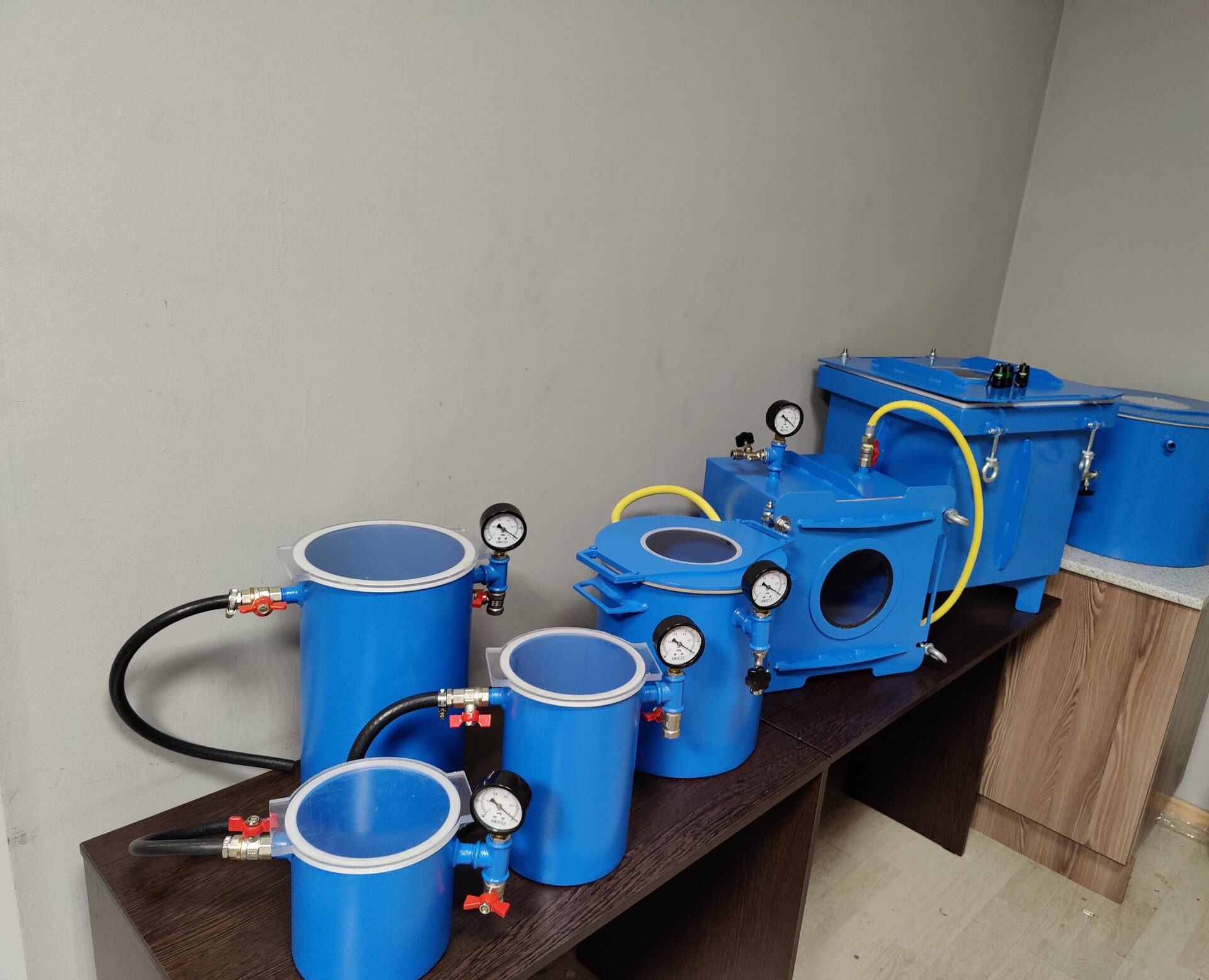 Готовые синие вакуумные камеры разных размеров с манометрами и шлангами, выставленные на столе в производственном помещении.