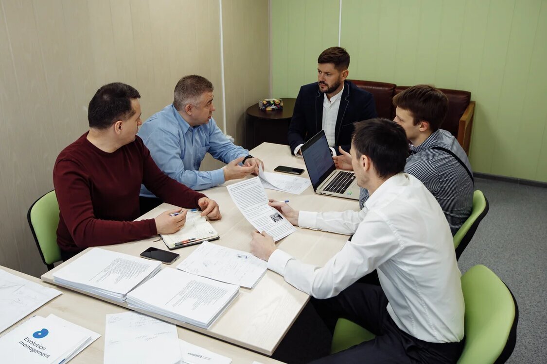 Команда обсуждает документы за столом с ноутбуком; Юрий в центре на дальней стороне стола