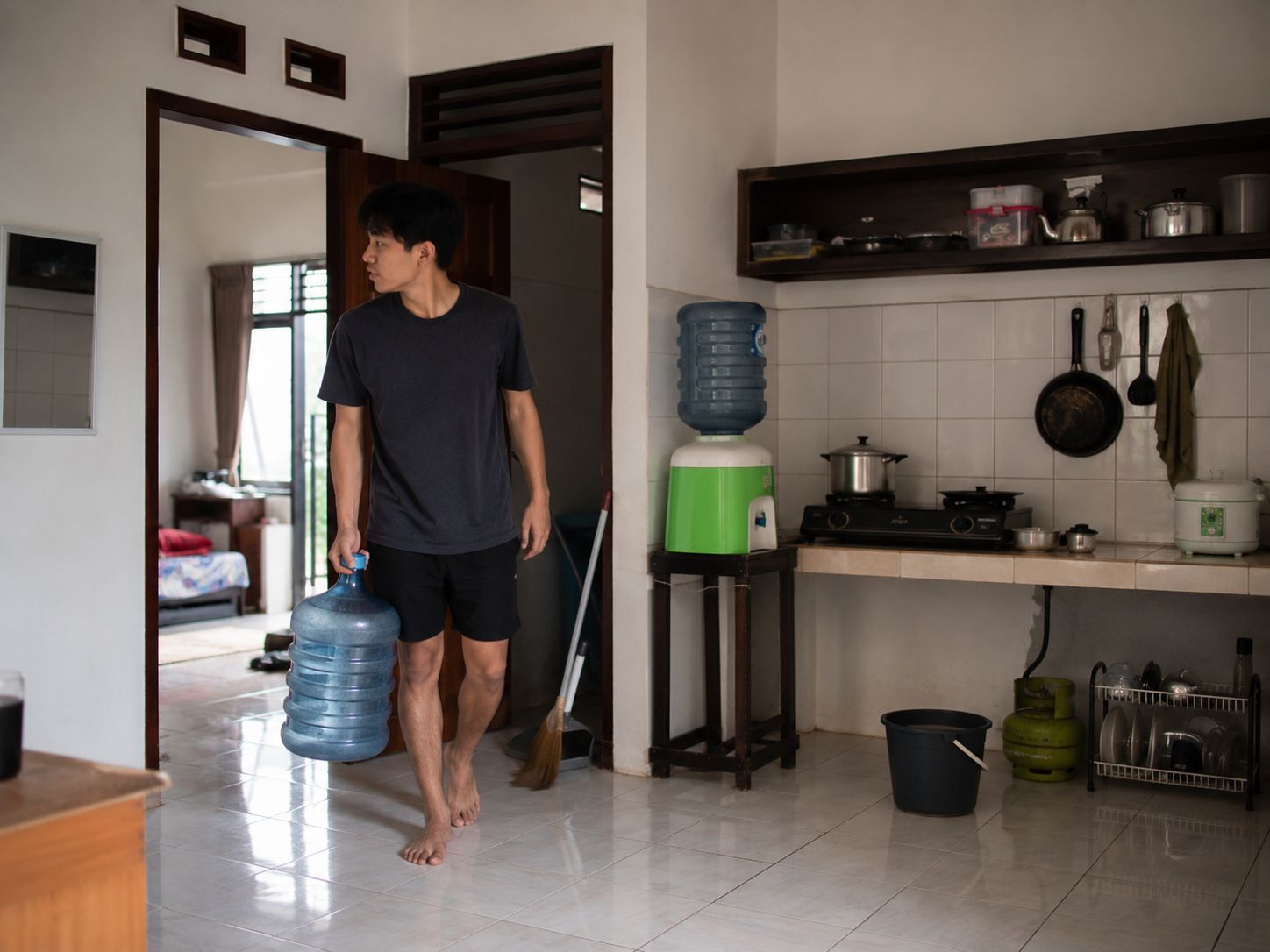 Penghuni kontrakan memegang galon kosong di dapur kecil dengan perlengkapan rumah sehari-hari
