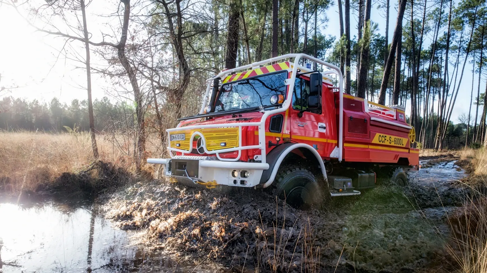 Красно-жёлтая пожарная машина Unimog едет по мутной воде в лесу
