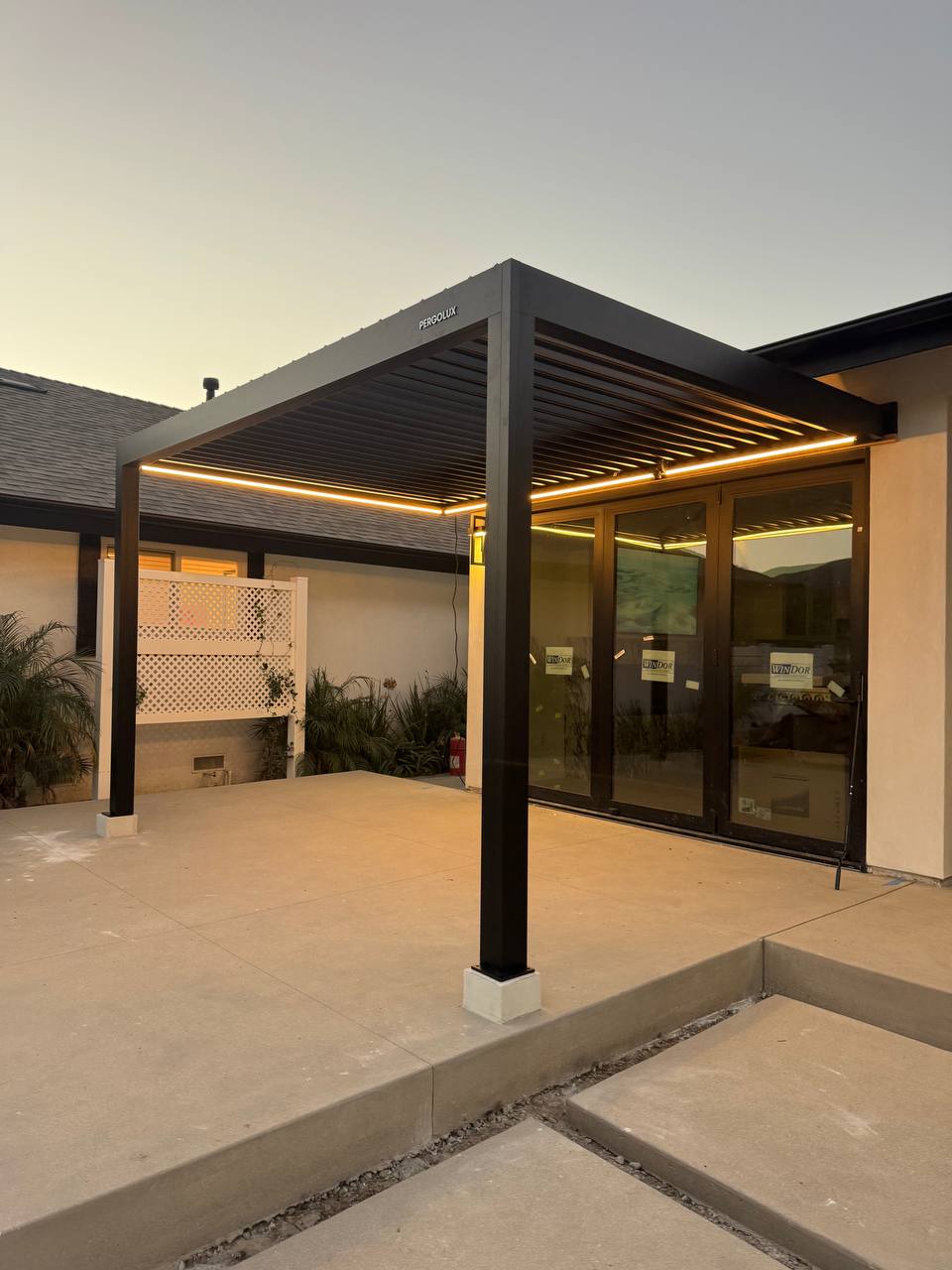Black pergola illuminated by warm LED strips beside the house at dusk.
