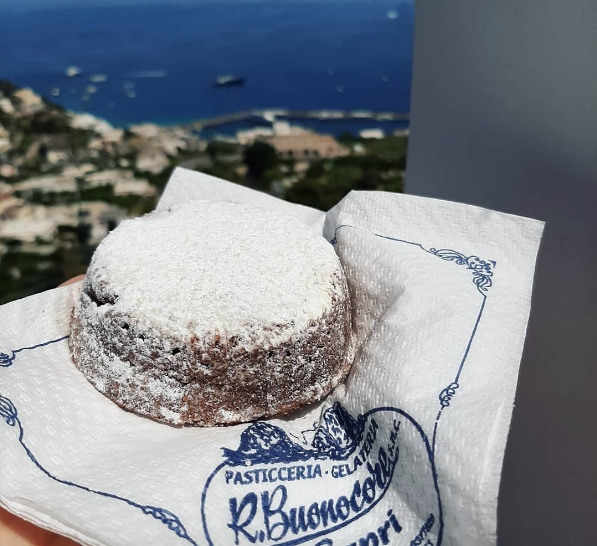 Маленький круглый торт Капрезе, обильно посыпанный сахарной пудрой, на фирменной салфетке кондитерской Buonocore на фоне панорамы острова Капри.