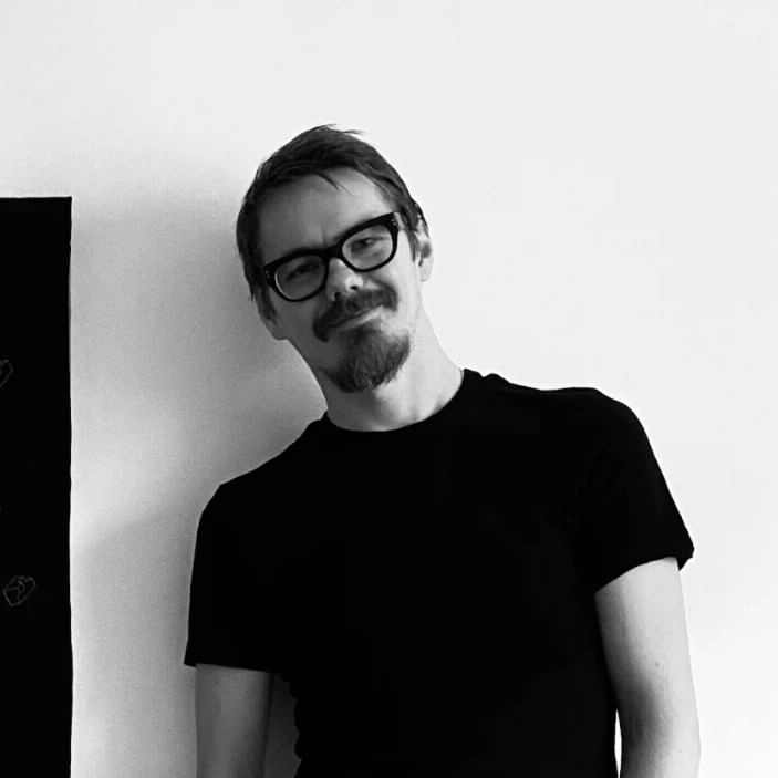 Монохромная портретная фотография мужчины в очках с темной оправой и небольшой бородкой, в черной футболке, на фоне белой стены с размытым темным пятном слева.