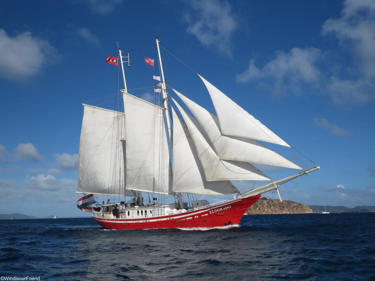 The red expedition schooner Eldorado sailing under full white sails on a deep-blue sea beneath a clear sky. The two-masted vessel designed for long-distance and polar expeditions glides through calm waters with a rocky island visible in the background.