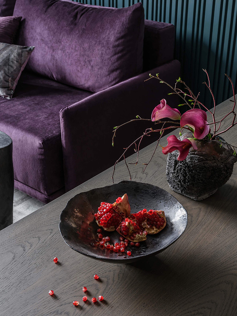 Дизайн интерьера в насыщенных тонах, деревянный стол, фиолетовый диван, зелёные стены насыщенного цвета. Гранат и розовые цветы в вазе