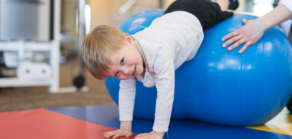Маленький мальчик на занятии у нейропсихолога тянется на синем мяче, смотря в камеру и улыбаясь. Его радостное выражение лица и активное участие в упражнении передают атмосферу доверия и интереса к процессу. Синий мяч, используемый для развития координации и motor skills, становится важным элементом занятия. Этот момент идеально подходит для статей о нейропсихологических методиках, развитии детей, therapeutic играх или материалах, посвященных поддержке детей с особыми потребностями. Картинка вызывает теплые эмоции и напоминает о важности профессионального подхода и заботы в развитии каждого ребенка.