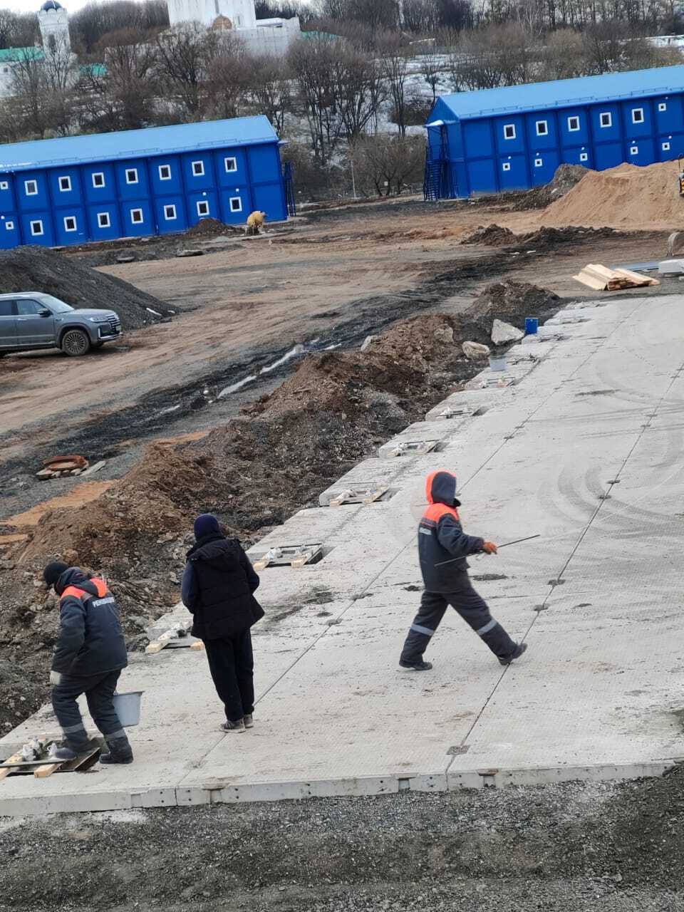 Рабочая бригада выполняет подготовку и заделку швов на площадке из плит ПАГ 14 ТУ на фоне синих модулей временного городка.