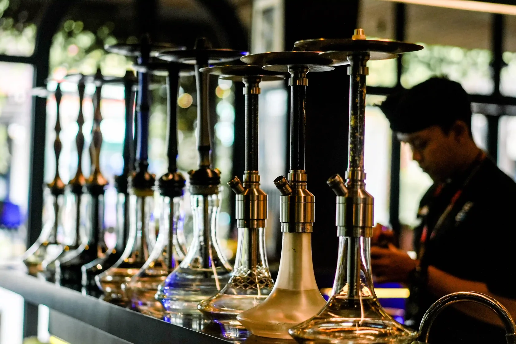 A stylish lineup of elegant glass hookah pipes sitting neatly on a lounge counter, ready for a relaxing session, with a staff member softly blurred in the background.