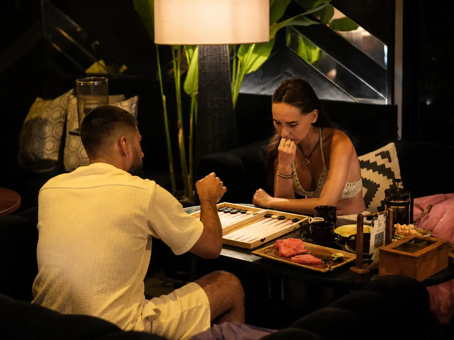 A couple in a relaxed, intimate lounge environment playing a game of Backgammon on a wooden board, with refreshments and tropical views in the background.