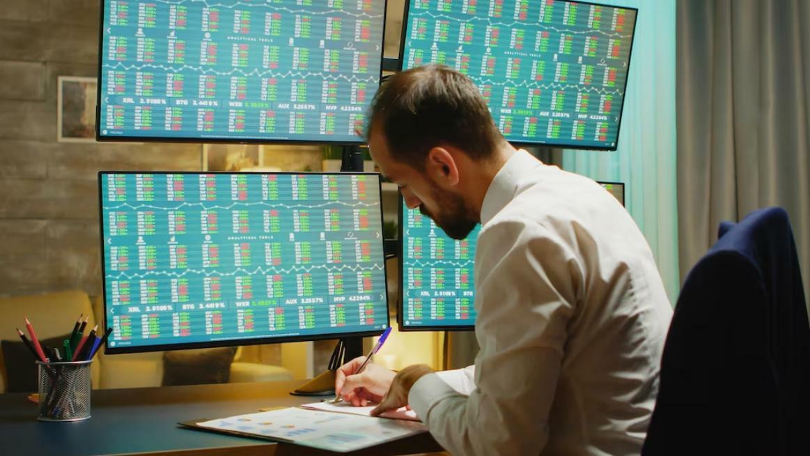 Un hombre sentado frente a cuatro monitores con gráficos de acciones, escribiendo en una hoja las señales de trading.