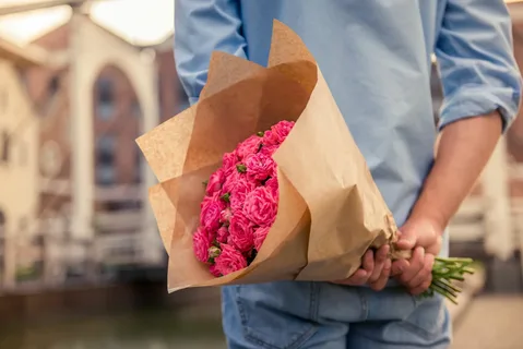 Мужчина денржит за спиной букет из розовых пионовидных роз, упакованный в крафт бумагу