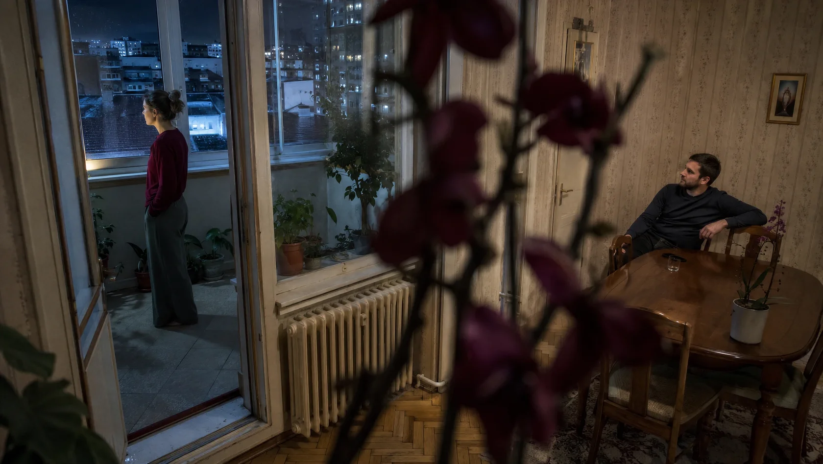 Женщина стоит на балконе, а мужчина сидит за столом в комнате после разговора, закончившегося молчанием.