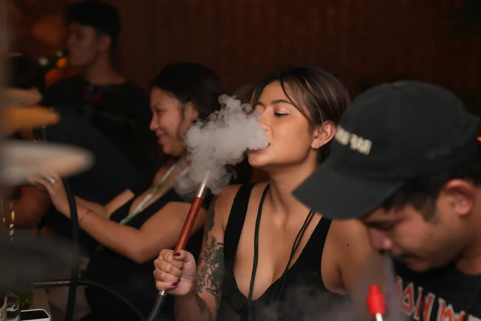 A young woman enjoying her first time trying hookah, exhaling a smooth cloud of smoke while relaxing with friends in a vibrant and cozy lounge setting.