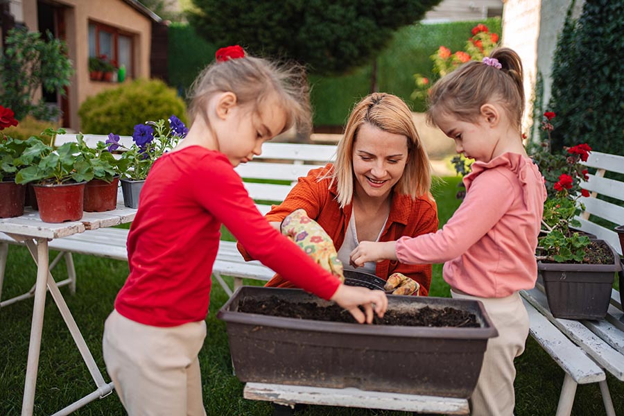 Woman gardening with two children, planting seeds in a container, surrounded by potted flowers, emphasizing beginner gardening and family involvement.
