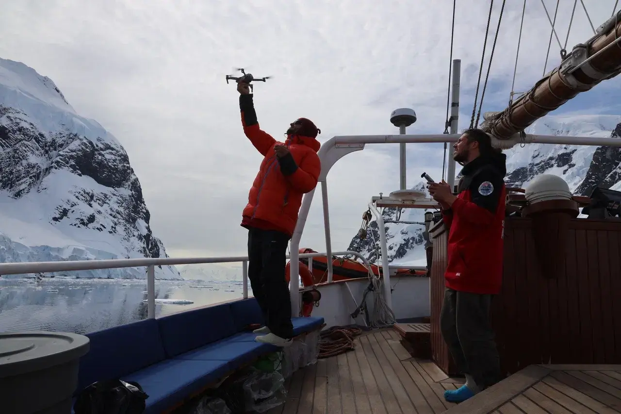 Antarctic expedition crew using a drone aboard a sailing yacht near glaciers and ice-covered peaks