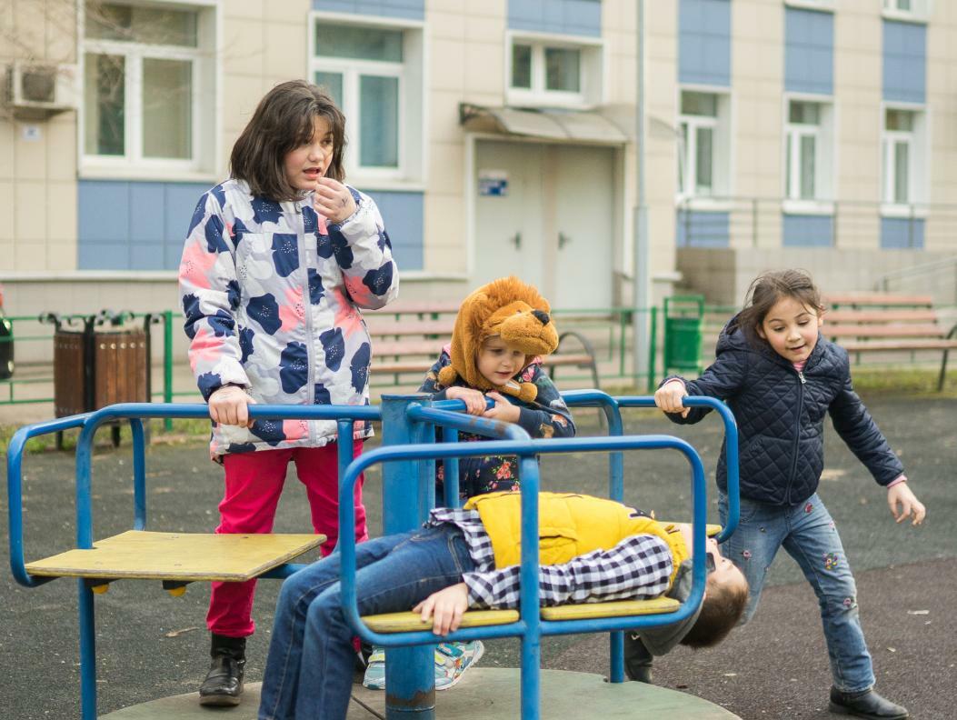 Ваня в белой футболке, зеленой куртке и синих джинсах стоит на фоне белого здания с большими окнами