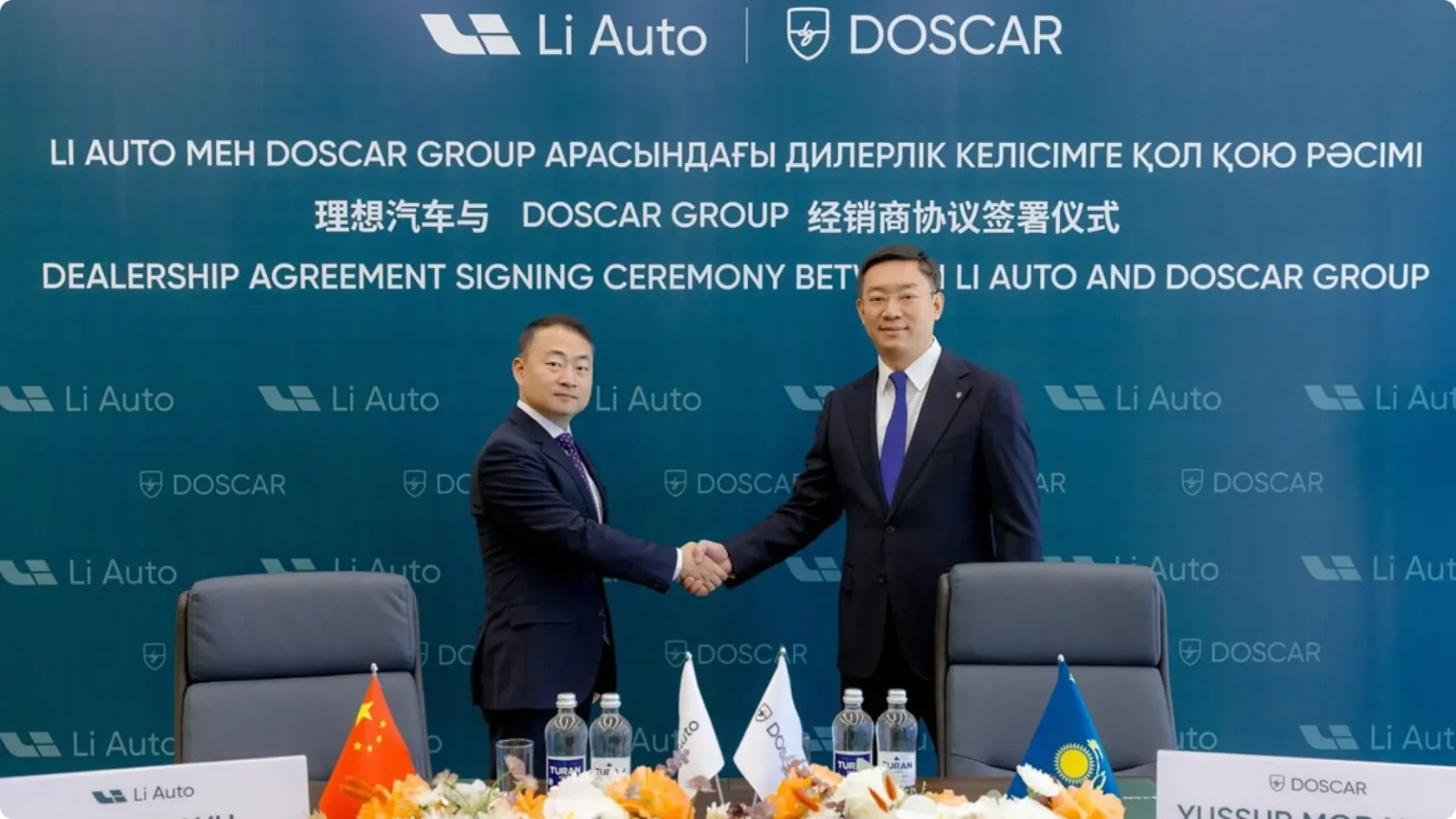 Церемония подписания дилерского соглашения между Li Auto и казахстанской группой DOSCAR: два представителя в деловых костюмах пожимают руки на фоне брендированного пресс-вола с флагами Китая и Казахстана.