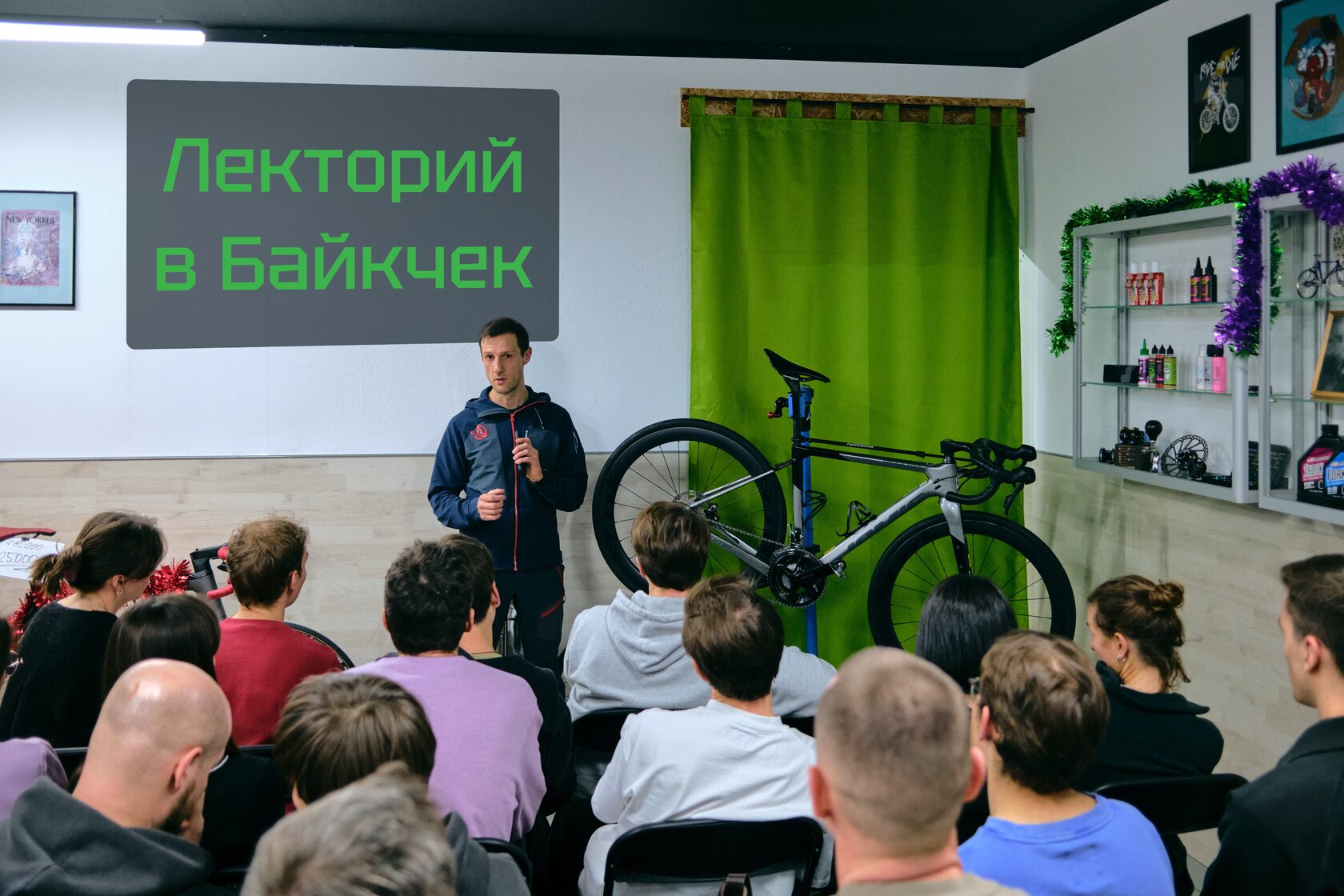 Ведущий выступает перед слушателями в веломастерской рядом с велосипедом на ремонтной стойке