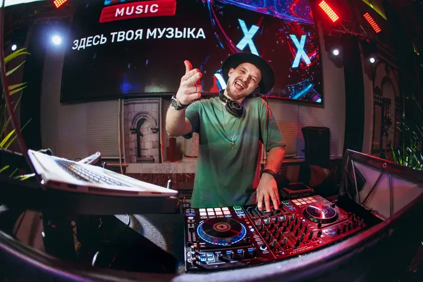 DJ ROZHKOV в зелёной футболке улыбается и показывает большой палец за диджейским контроллером.
