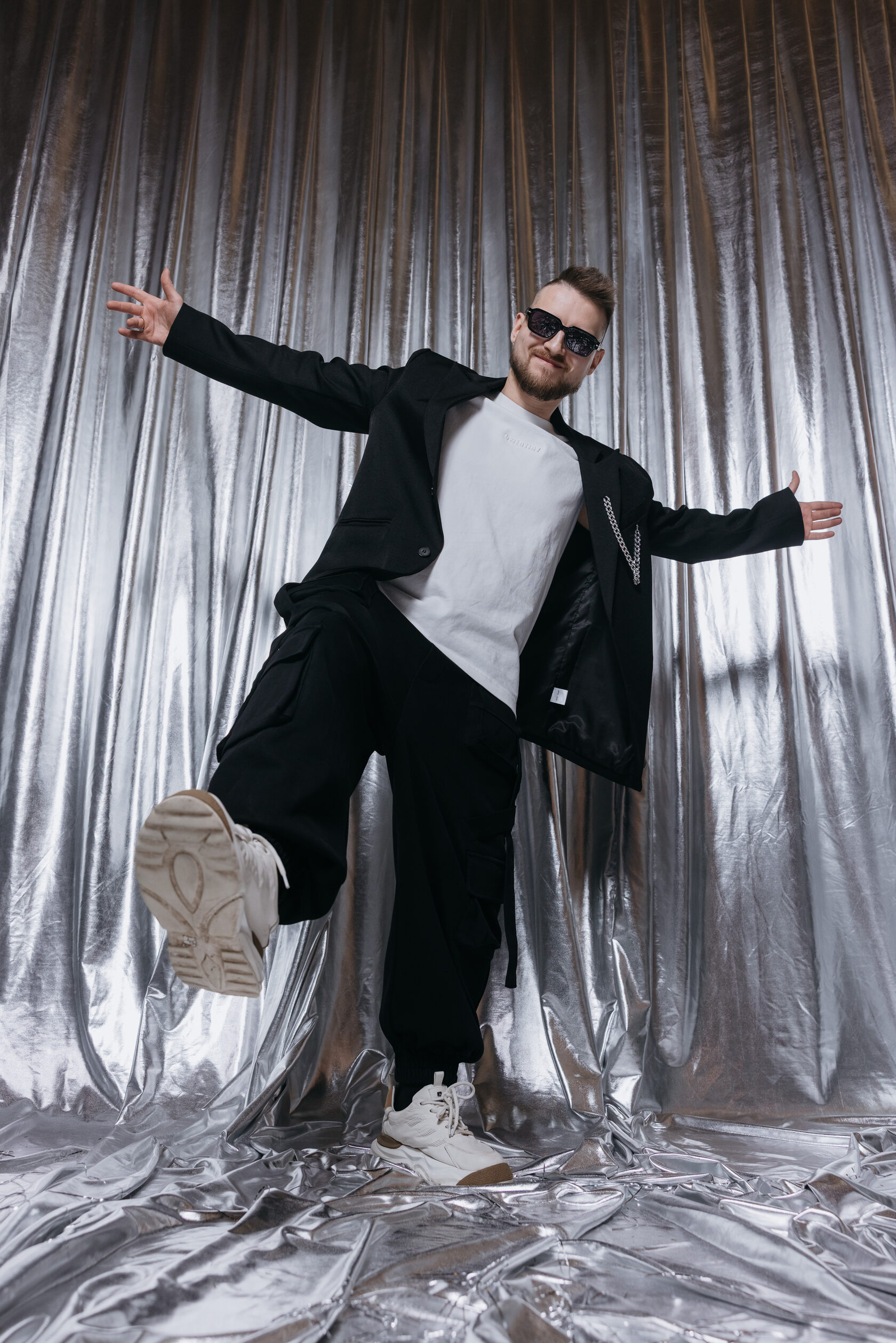 DJ ROZHKOV в чёрном пиджаке и очках позирует на фоне серебристого занавеса.