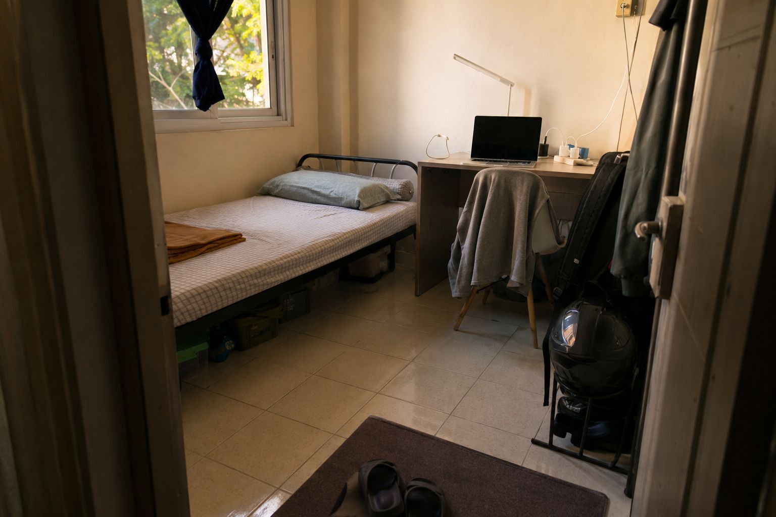 Kamar kos Indonesia dengan meja kerja, charger, pakaian di kursi, dan sandal dekat pintu