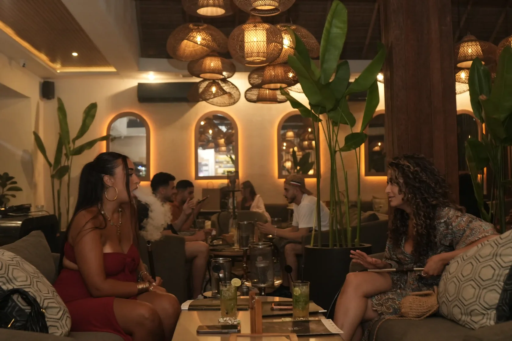 Two female friends relaxing on a cozy sofa at Eden Hookah Club Canggu, with one blowing thick shisha smoke while enjoying a great conversation.