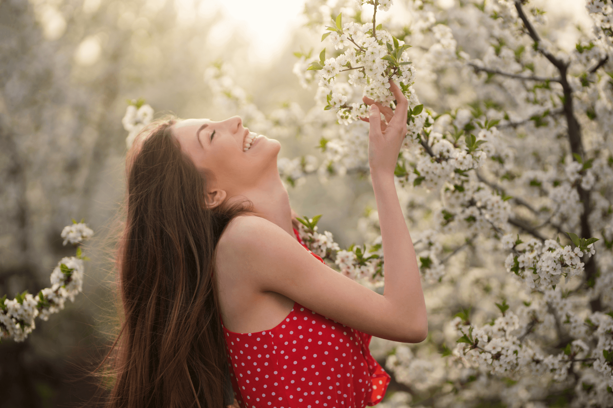 Frau ist von blühenden Bäumen umgeben, berührt eine weiße Baumblüte und lächelt dabei in Richtung Himmel