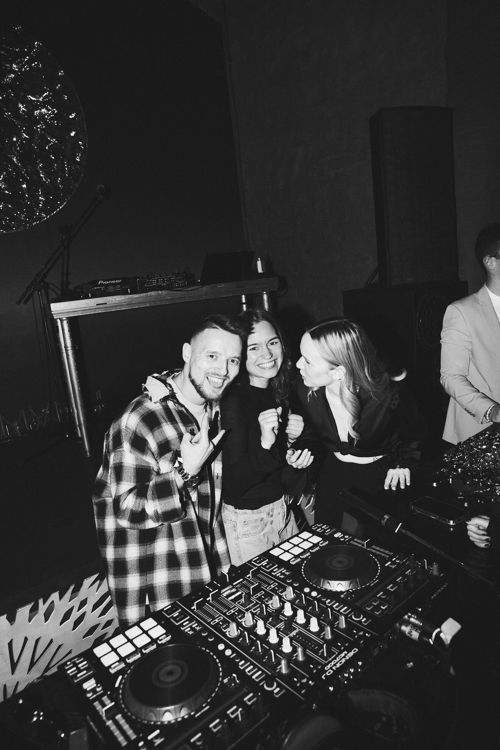Чёрно-белый кадр DJ ROZHKOV за диджейским пультом рядом с улыбающимися гостями.