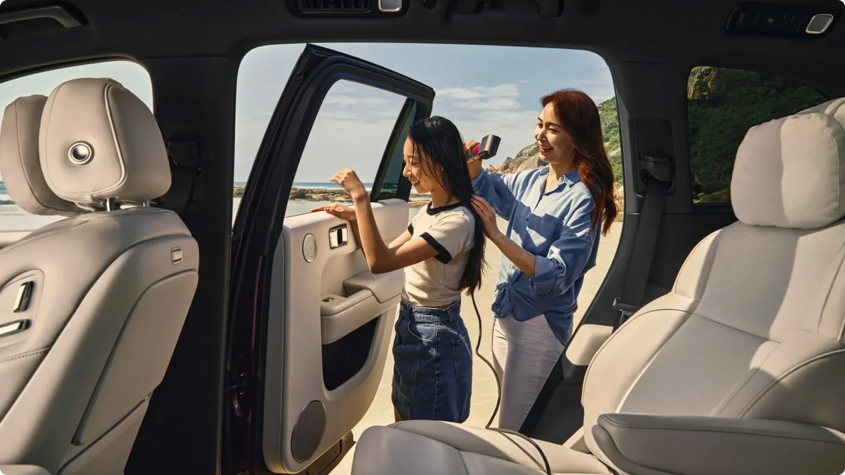 Использование розеток в салоне Li Auto L9: женщина сушит волосы девочке феном, подключенным к автомобилю на берегу моря.