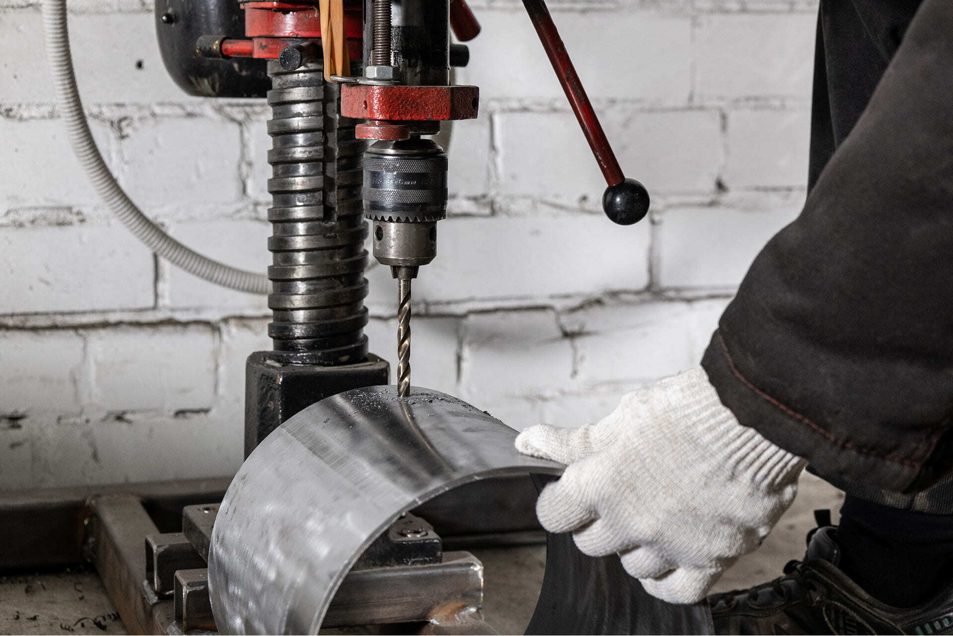Сверлильный станок в работе: мастер в перчатках делает отверстие в изогнутой металлической детали корпуса камеры