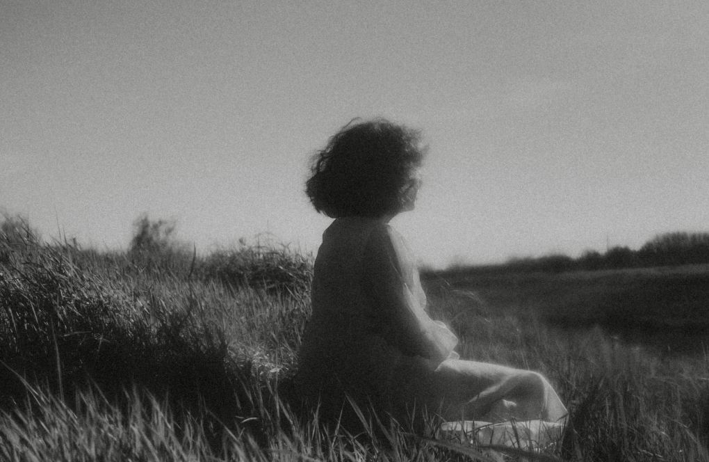 Девушка с короткими волосами в легком белом платье сидит в поле на траве