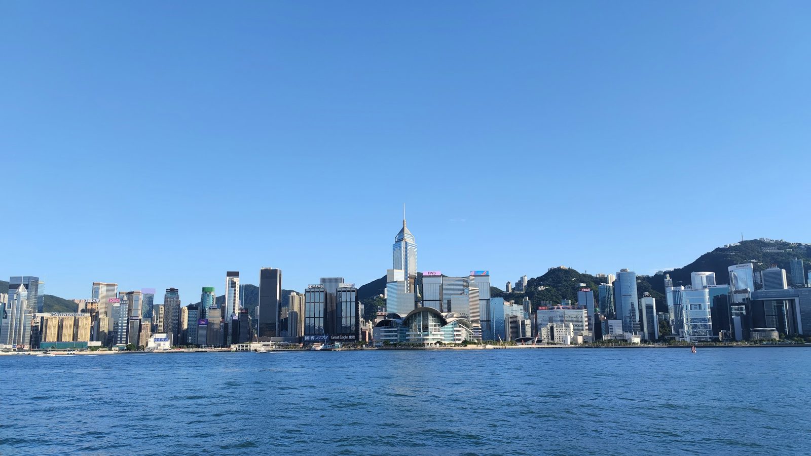 Hong Kong Island skyline across Victoria Harbour on a clear day, with the Convention and Exhibition Centre on the waterfront and green hills behind