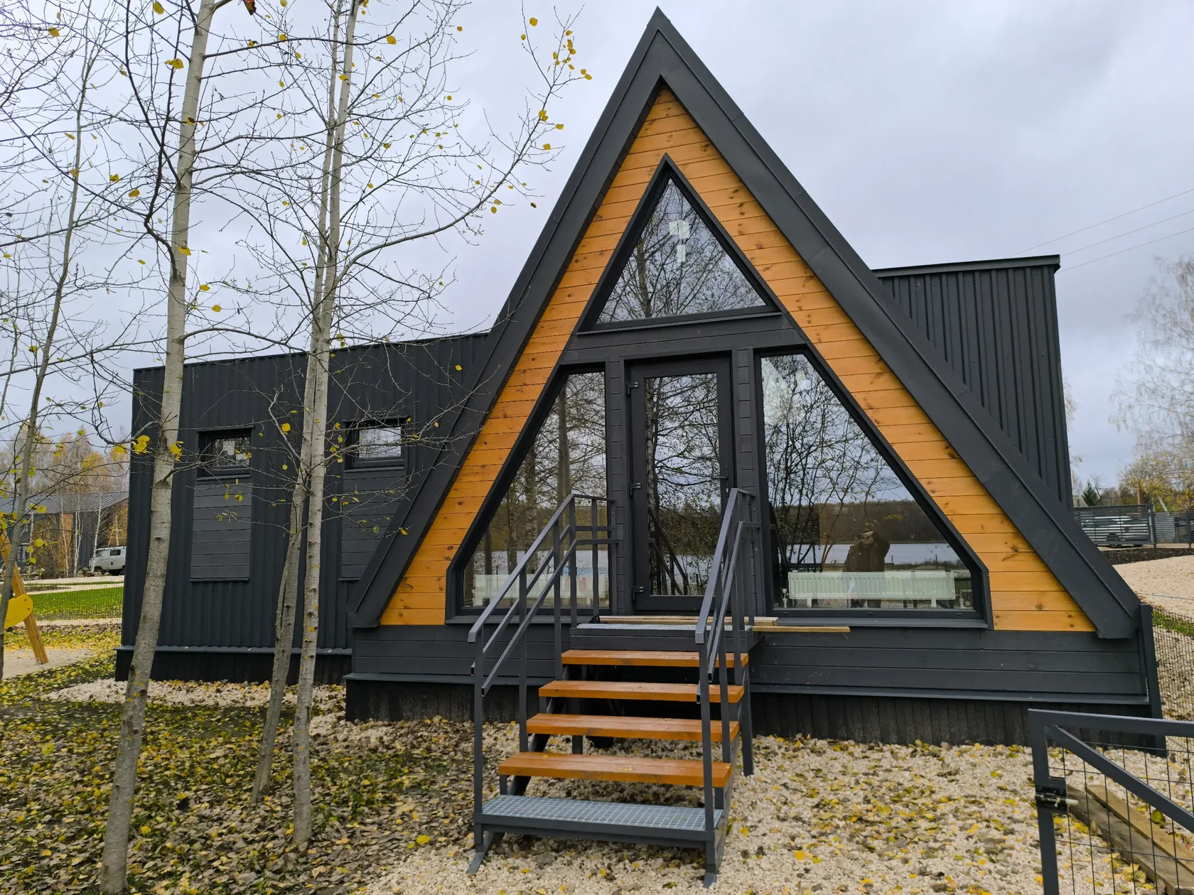Треугольный дом A-frame с черной фальцевой кровлей и большим треугольным окном на фасаде.