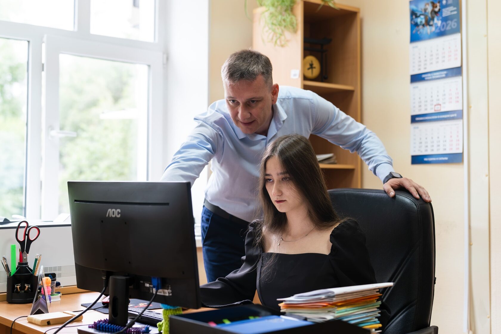 Девушка-подросток сосредоточенно работает за компьютером в офисе, пока мужчина-наставник в голубой рубашке указывает на экран и подсказывает алгоритм действий.