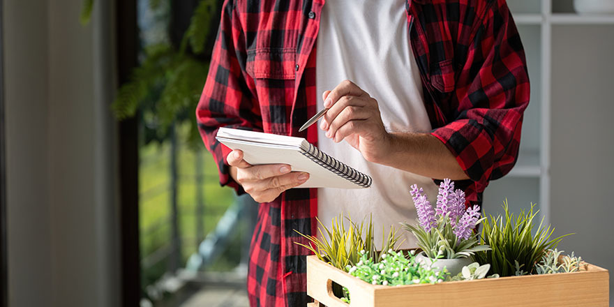 Person in a red plaid shirt holding a notebook and pen, surrounded by potted plants, illustrating gardening planning and organization.
