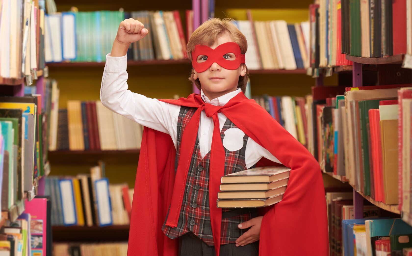 a white boy in a library, wearing a cape and mask with his arm in the air like a super hero and holding books in the other