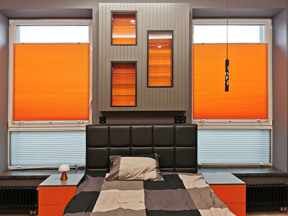 кровать с чёрным кожаным изголовьем, оранжевые тумбы, оранжевое плиссе, серый подвесной стеллаж с оранжевыми нишами