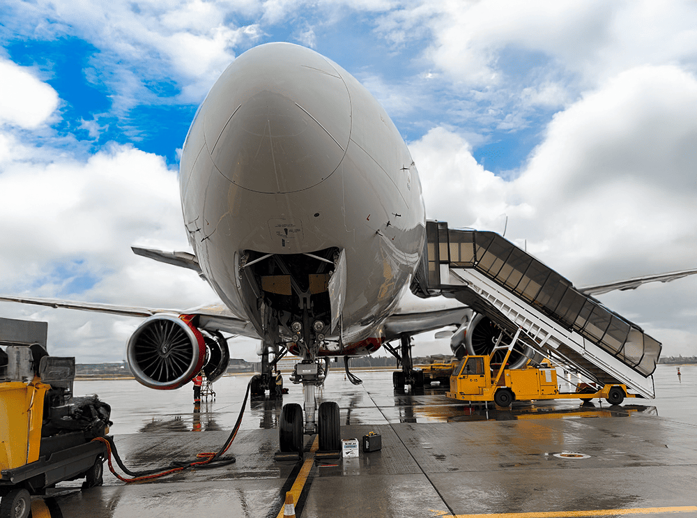 **Alt:** Самолет стоит на взлетно-посадочной полосе с носом, направленным к камере. Самолет находится в процессе техобслуживания или подготовки к вылету. Видна передняя стойка шасси, а грузовые двери открыты. Сбоку самолета находится мобильный трап, обесп