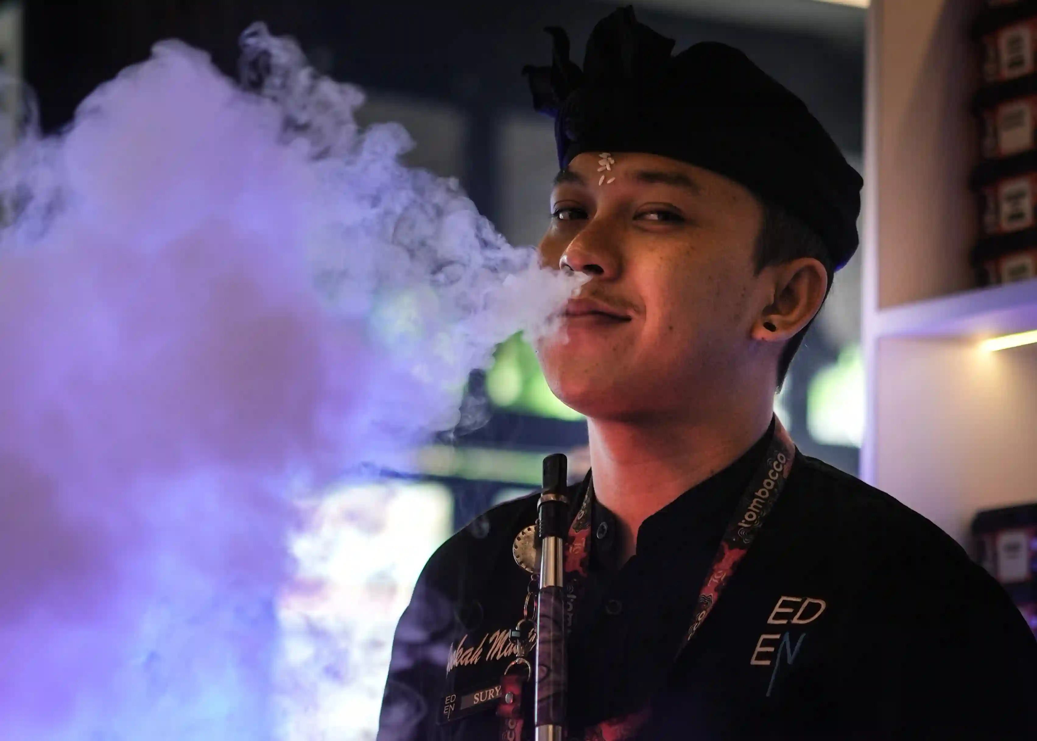 A friendly shisha master at Eden Hookah Club, wearing a traditional Balinese udeng, smiles warmly at the camera while blowing a smooth cloud of smoke.