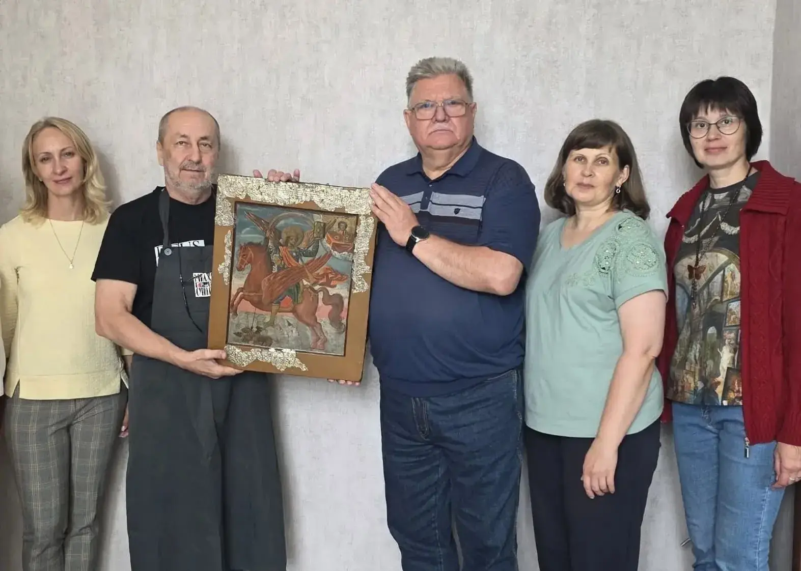 На фото — группа из пяти человек (три женщины и двое мужчин) позируют на фоне светлой стены. В центре двое мужчин держат икону в деревянной раме с декоративной окантовкой: на ней изображён святой Архангел Михаил. Остальные участники стоят по бокам, выраже